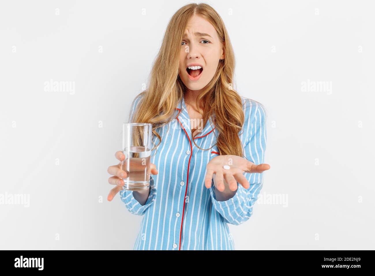 Portrait d'une jeune femme triste en pyjama sensation de douleur et tenant une pilule et un verre d'eau debout sur fond blanc Banque D'Images