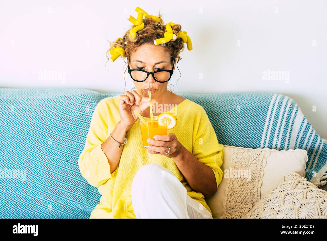 Portrait de couleur jaune de la belle femme de race blanche, jeune adulte, qui boit jus d'orange sain à la maison avec des curlers et un canapé bleu en arrière-plan - conce Banque D'Images