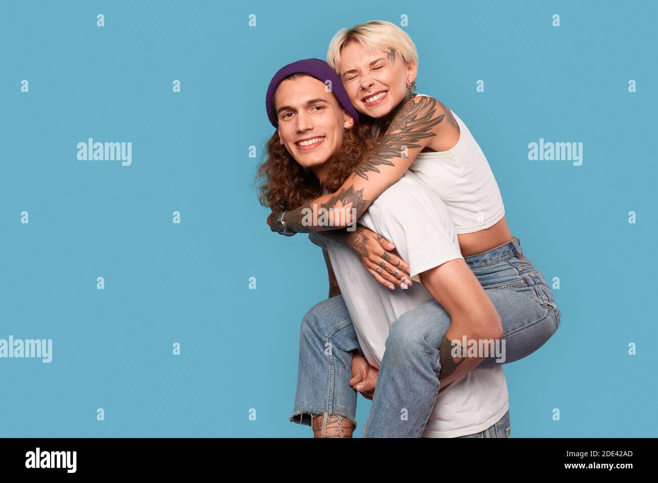 Style libre. Jeune couple tatoué debout isolé sur la petite amie bleue à cheval le dos de petit ami embrassant sourire heureux Banque D'Images