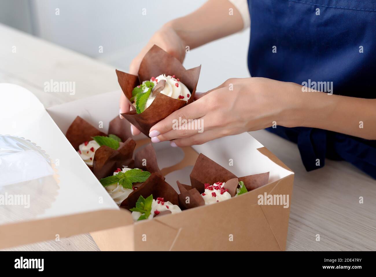 Vue rapprochée des mains de la confiserie ou du boulanger emballant des muffins savoureux décorés avec du plateau de crème à la boîte de livraison. Petits gâteaux prêts avec baies et Banque D'Images