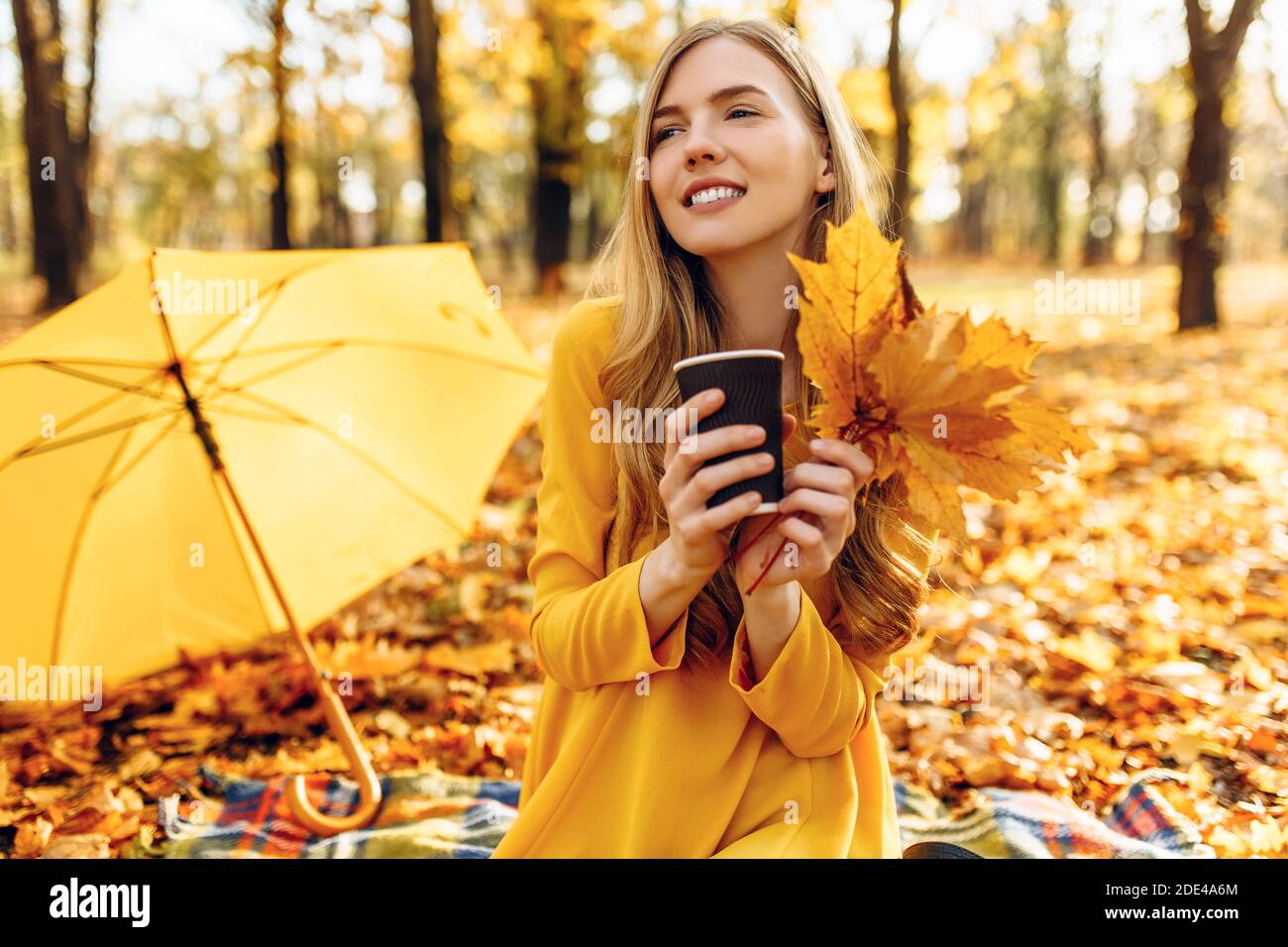 Belle jeune fille dans le parc d'automne assis sur une couverture, tenant une boisson chaude, appréciant le temps ensoleillé chaud Banque D'Images