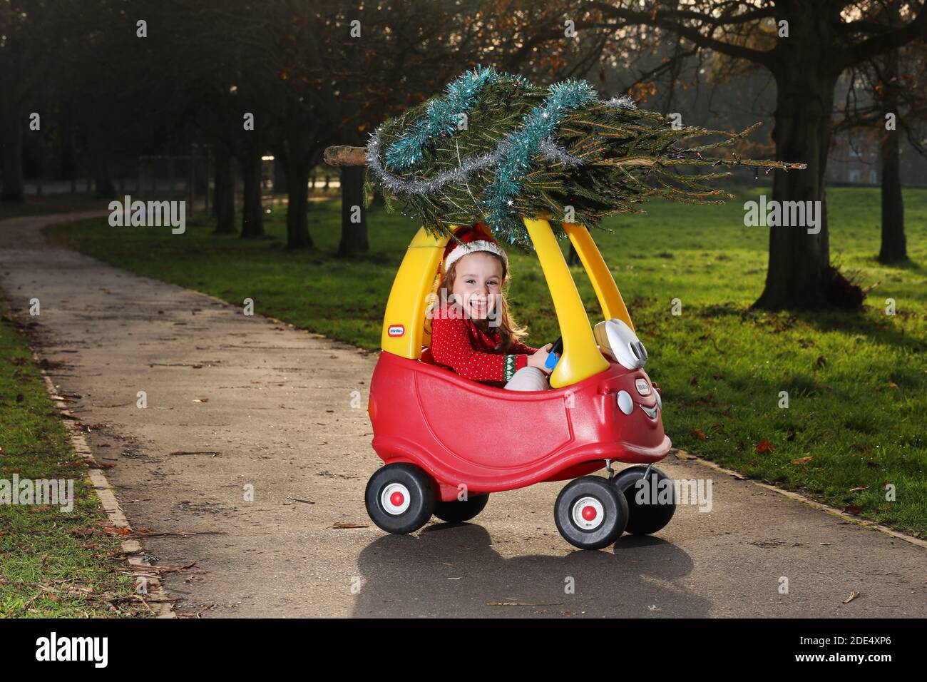 Jolie fille de cinq ans, Florence, représentée sous la forme d'un Elf de Noël et présentant un petit arbre de Noël sur sa voiture rouge classique Little Tikes, au Royaume-Uni. Banque D'Images