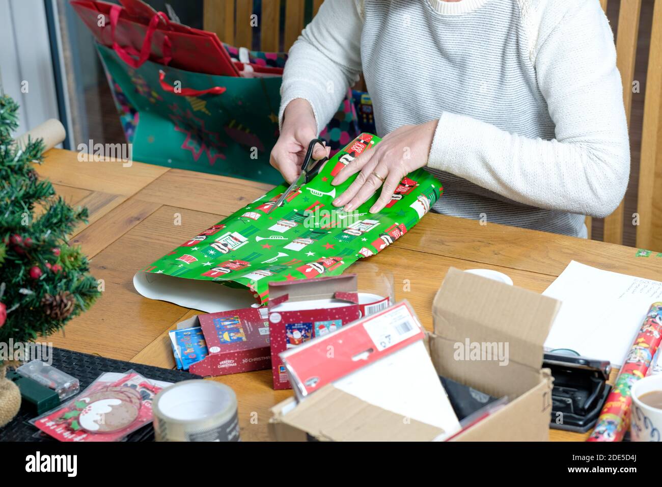 Une femme, assise à une table, enveloppe un cadeau de noël dans un papier cadeau de noël aux couleurs vives. Elle tonde le papier à la taille avec des ciseaux Banque D'Images