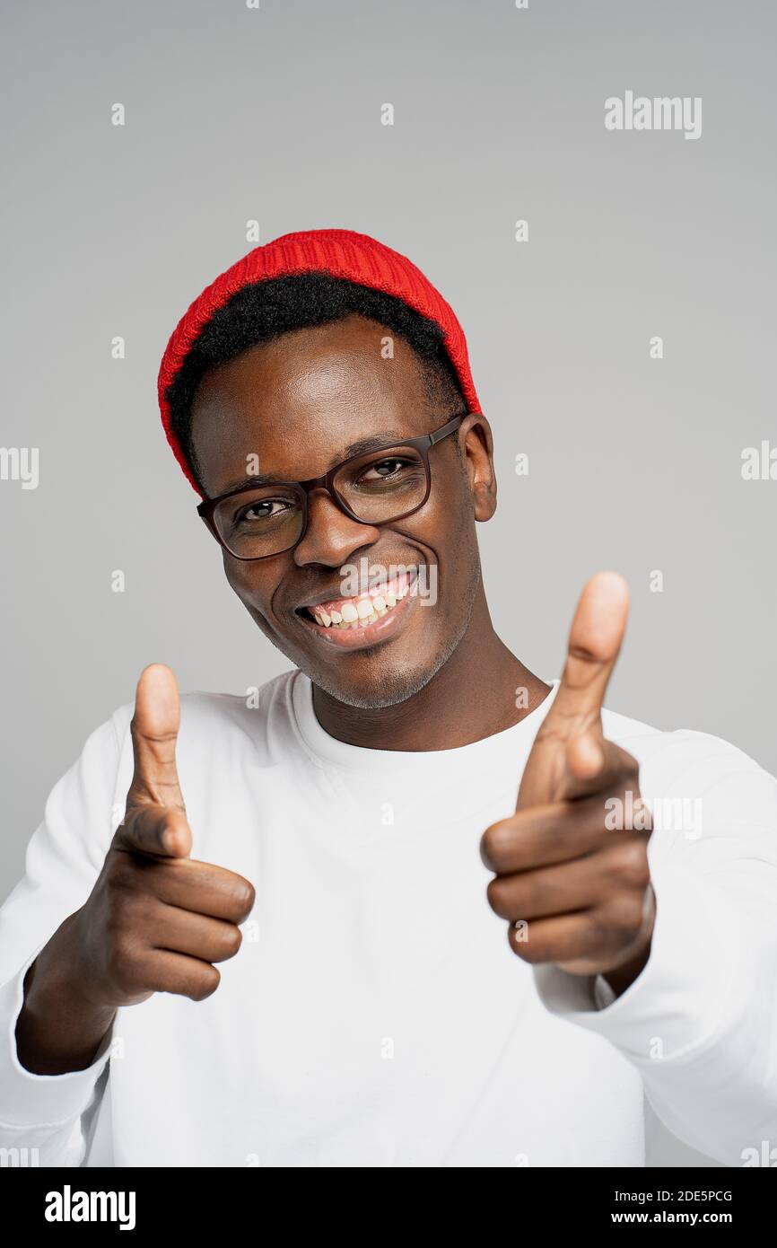 Hé, vous! Joyeux positif jeune Afro américain homme porter un chapeau rouge dans bonne humeur souriant large, pointant les doigts vers l'appareil photo tout en se tenant contre studi Banque D'Images