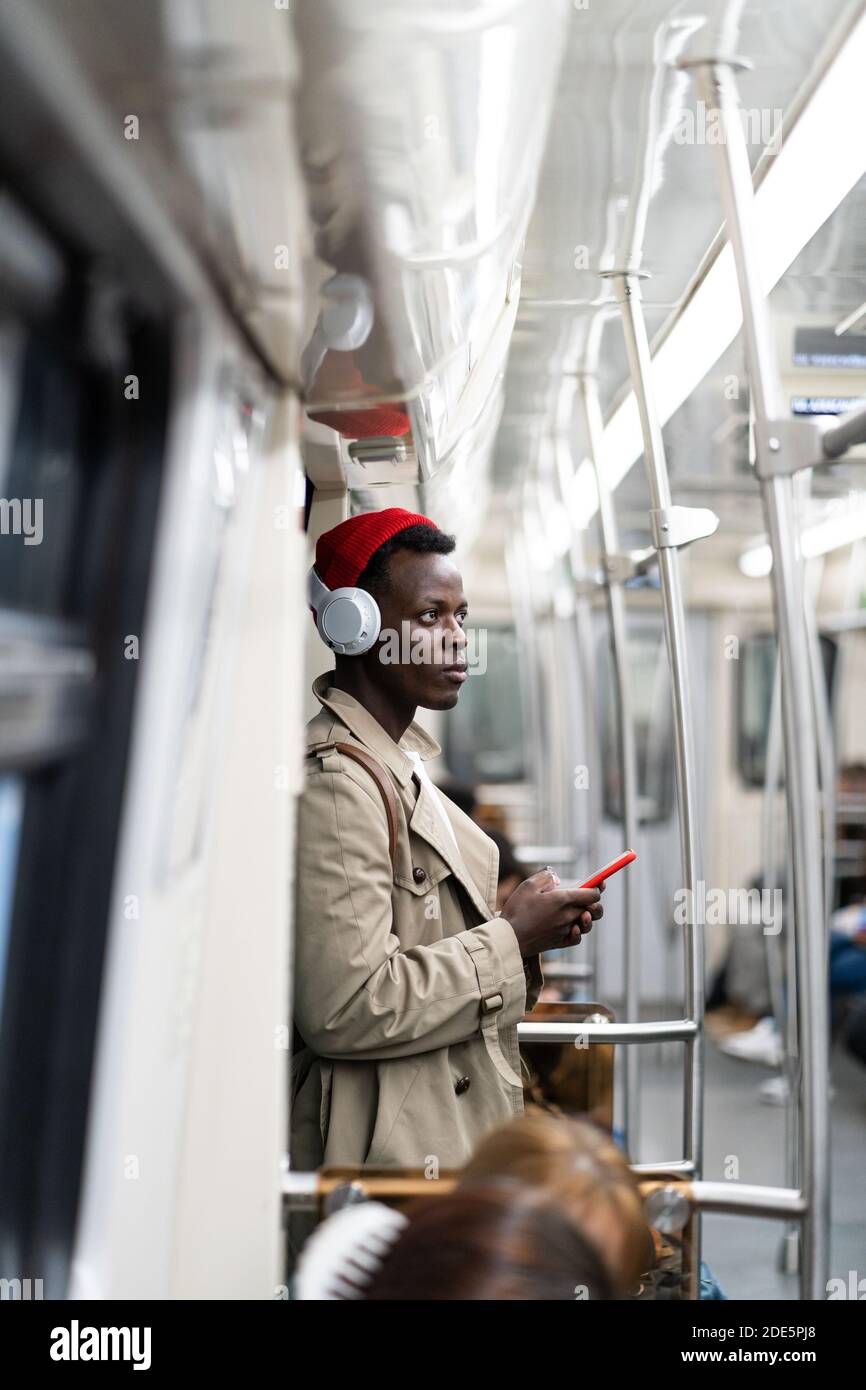 Un homme de passager afro-américain en chapeau rouge, un trench coat stand dans le métro, en utilisant un smartphone mobile, écoute de la musique avec des écouteurs sans fil dans publ Banque D'Images