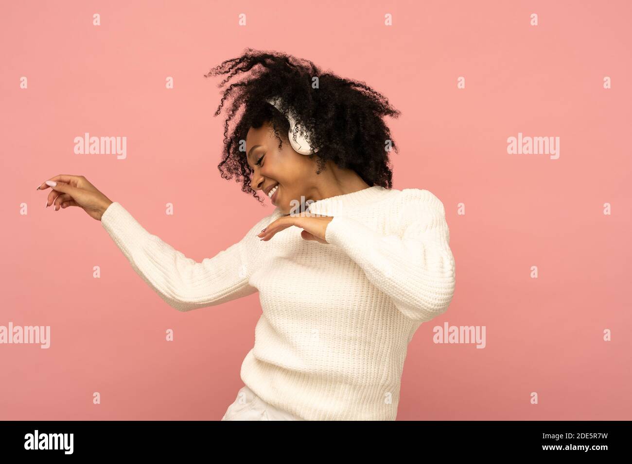 Femme biraciale active dans un chandail blanc chantent, porte des écouteurs sans fil, a une bonne humeur, isolé sur fond rose. Joyeux curly foncé à la peau Banque D'Images