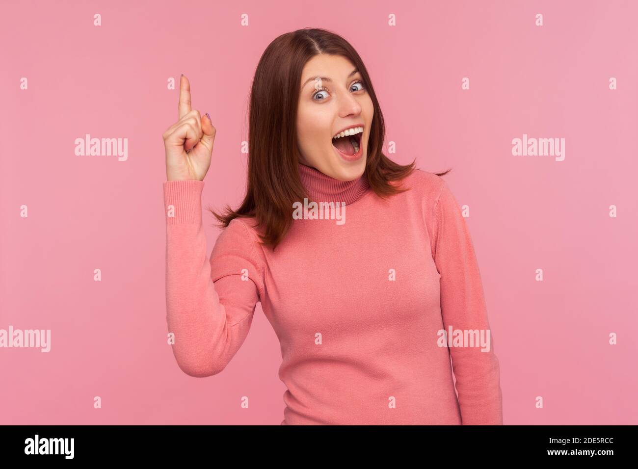 Une femme excitée et inspirée avec des cheveux bruns pointant vers le haut en regardant l'appareil photo avec un sourire crasseux, heureuse avec une nouvelle idée géniale, trouver la solution. Studio intérieur Banque D'Images