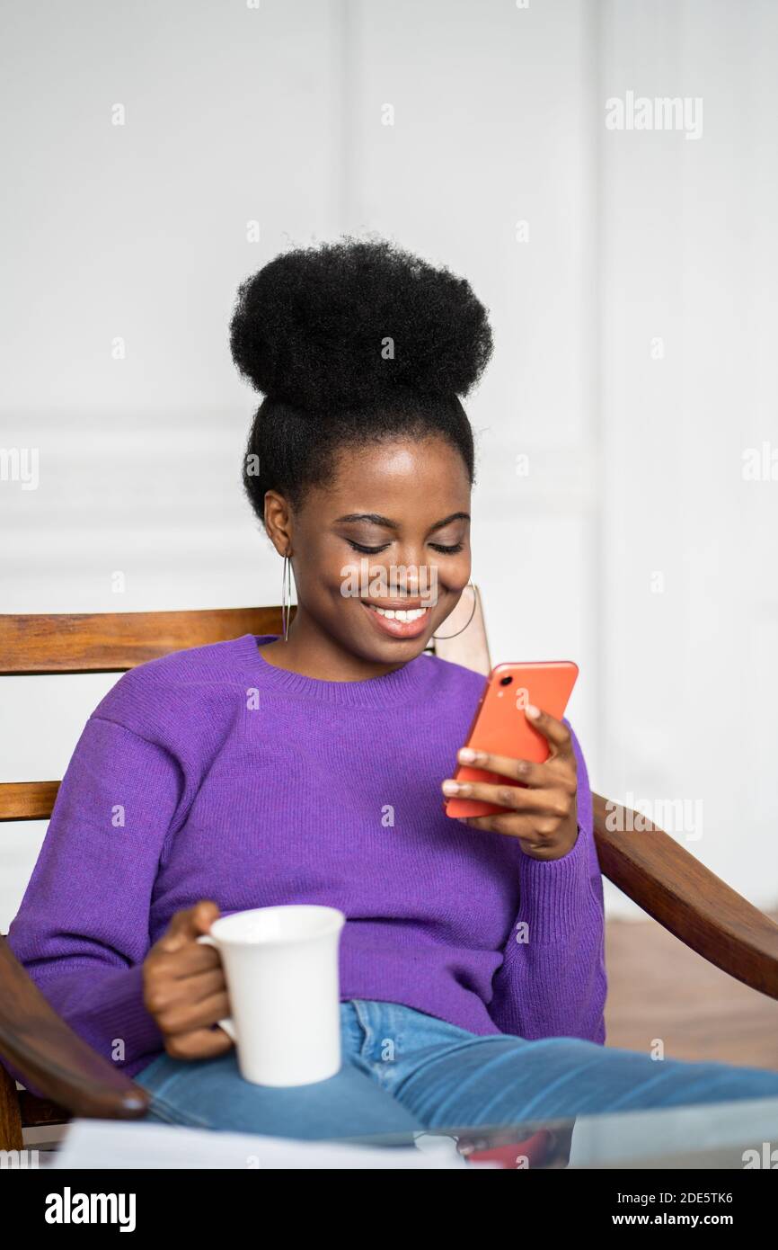 Gros plan d'Afro-américaine femme millénaire porter un chandail violet assis dans un fauteuil, en utilisant un smartphone mobile, en discutant dans les réseaux sociaux, en buvant te Banque D'Images