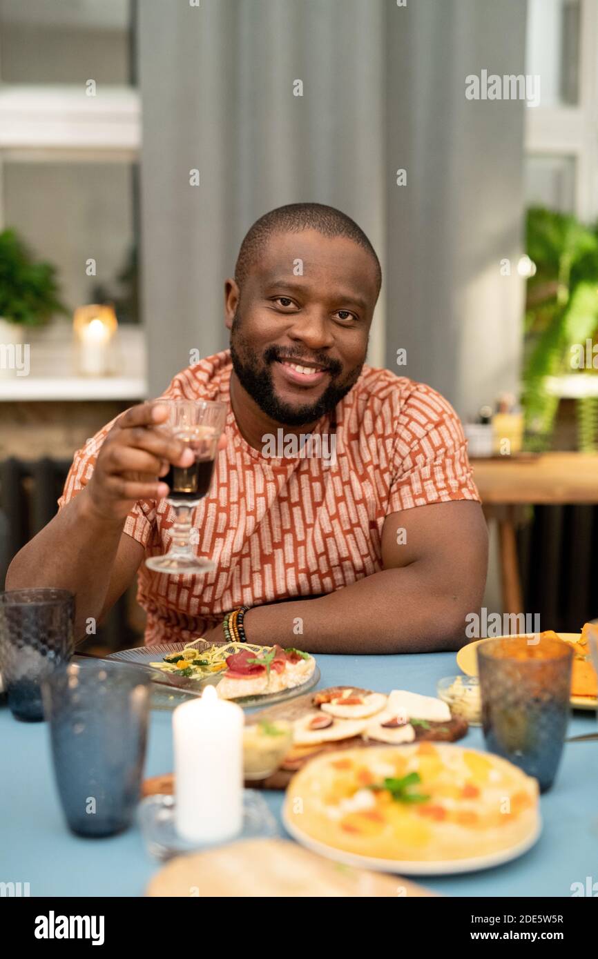 Joyeux jeune Africain qui applaudisse avec un verre de rouge vin devant l'appareil photo pendant que vous êtes assis près de l'hôtel, servi de fête Table pendant le dîner de Noël Banque D'Images