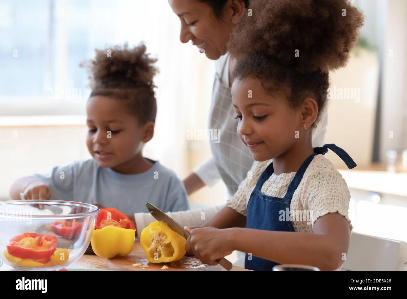 Une mère d'accueil noire souriante regarde les enfants adoptés cuisiner des plats de légumes Banque D'Images