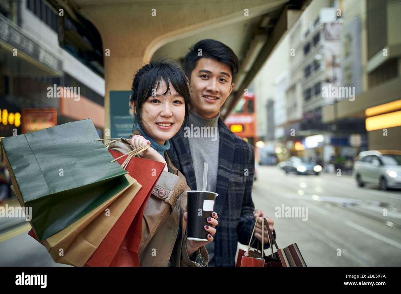 un jeune couple asiatique heureux attend le bus dans une ville moderne avec des sacs à provisions à portée de main Banque D'Images