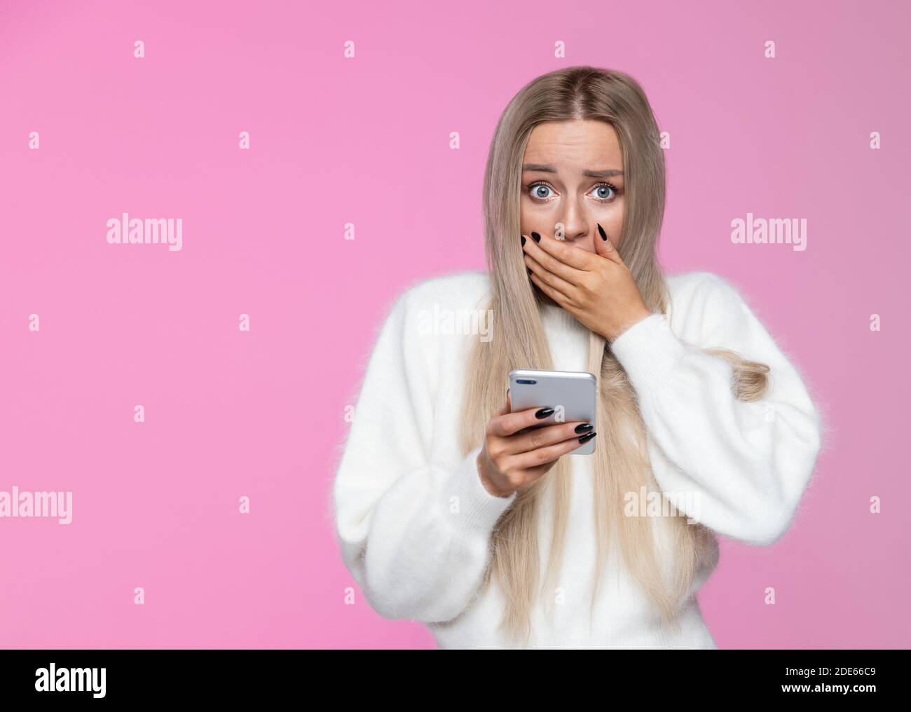 Une jeune femme blonde inquiète couvre la bouche en état de choc, tenant le téléphone portable, regardant la caméra, réaction surprise sur son visage, isolée sur fond rose Banque D'Images