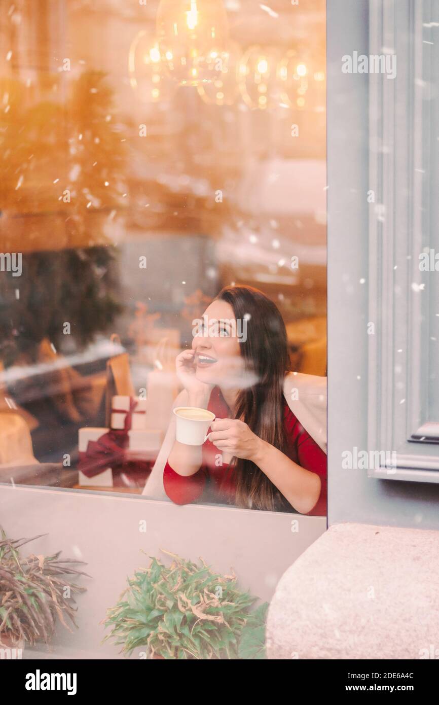 Jeune femme attrayante et gaie buvez le café du matin et regardez dans la fenêtre sur un steet enneigé. Bonne fille élégante et belle pause-café dans le restaurant du centre commercial Banque D'Images