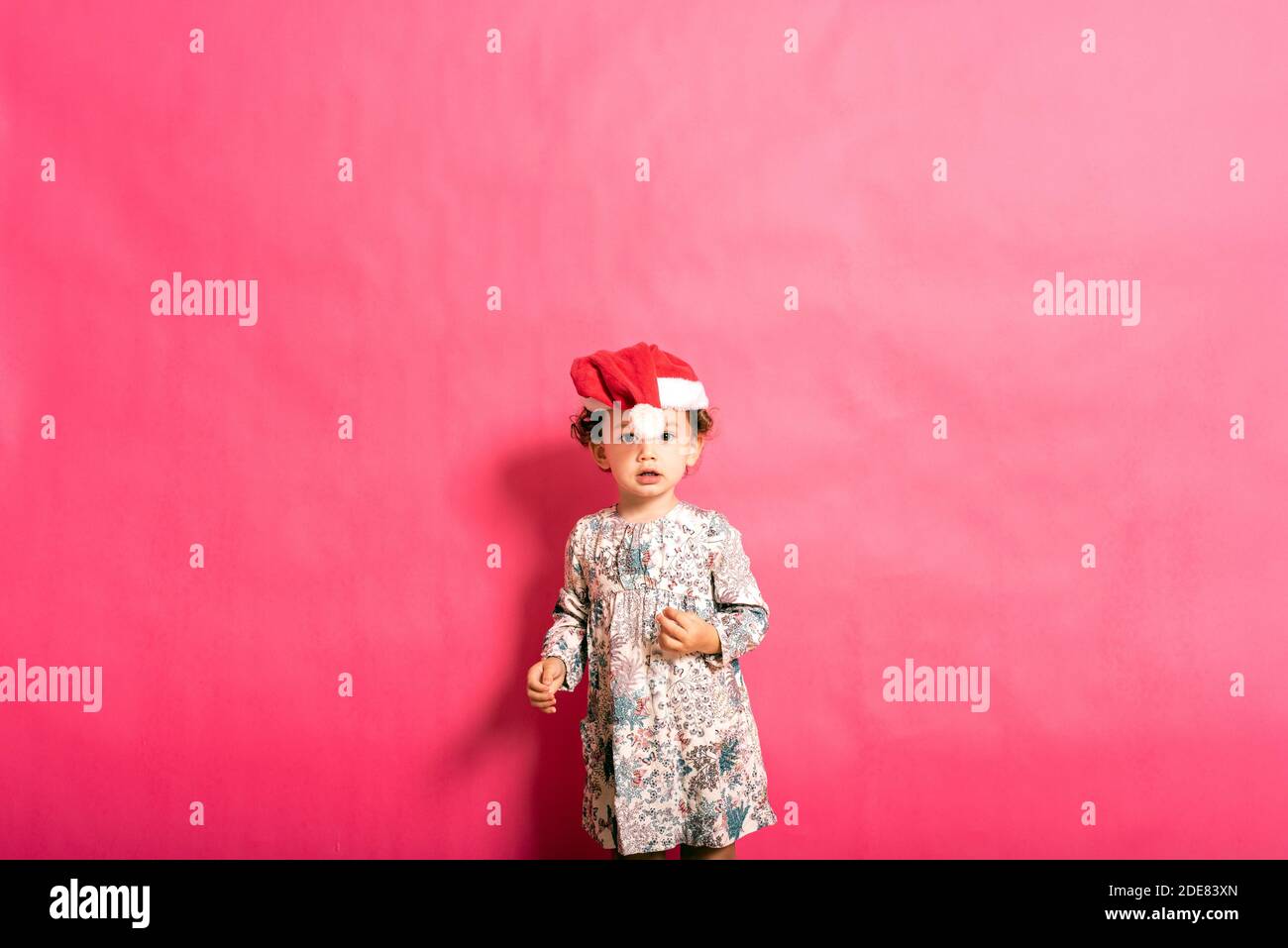 Bébé de deux ans avec un chapeau de père noël mal placé et un aspect drôle, isolé sur fond rose et espace de copie. Banque D'Images