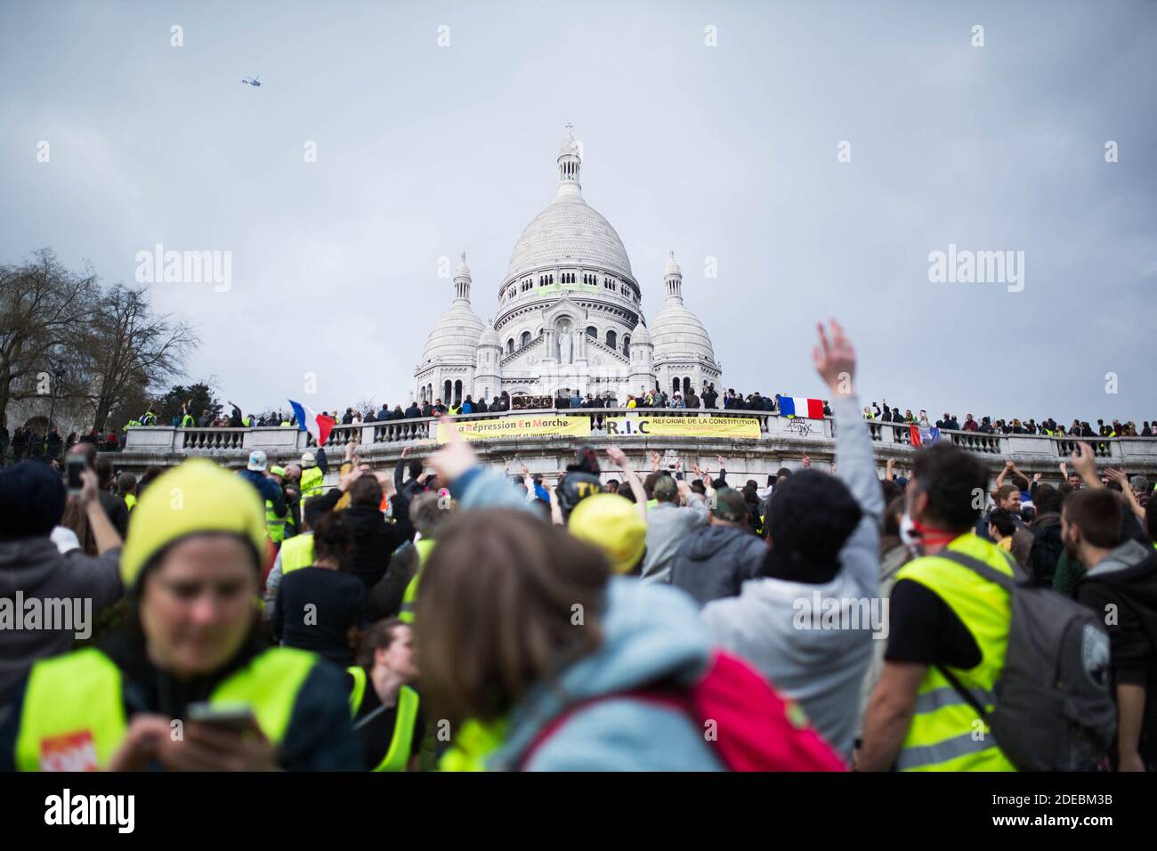 Les manifestants de la Yellow Vest se rassemblent devant la basilique du Sacré-cœur à Montmartre et regardent les manifestants qui fixent un panneau jaune sur le Sacré-cœur à côté d'un hélicoptère de police, à Paris le 23 mars 2019, lors d'une manifestation antigouvernementale organisée par le mouvement de la « Yellow Vest » (gilets jaunes). Les manifestants ont de nouveau frappé les rues de la ville française le 23 mars, pour une 19e semaine consécutive de protestation nationale ( Loi 19 ) contre la politique du président français et son style de gouvernement descendant, le coût élevé de la vie, les réformes fiscales gouvernementales et pour plus de "justice sociale et économique." photo de Raphael LAF Banque D'Images