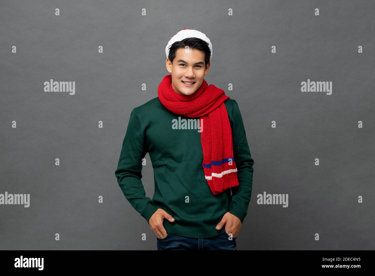 Jeune homme asiatique beau portant des couleurs rouges et vertes de Noël tenue en studio avec les mains dans la poche Banque D'Images