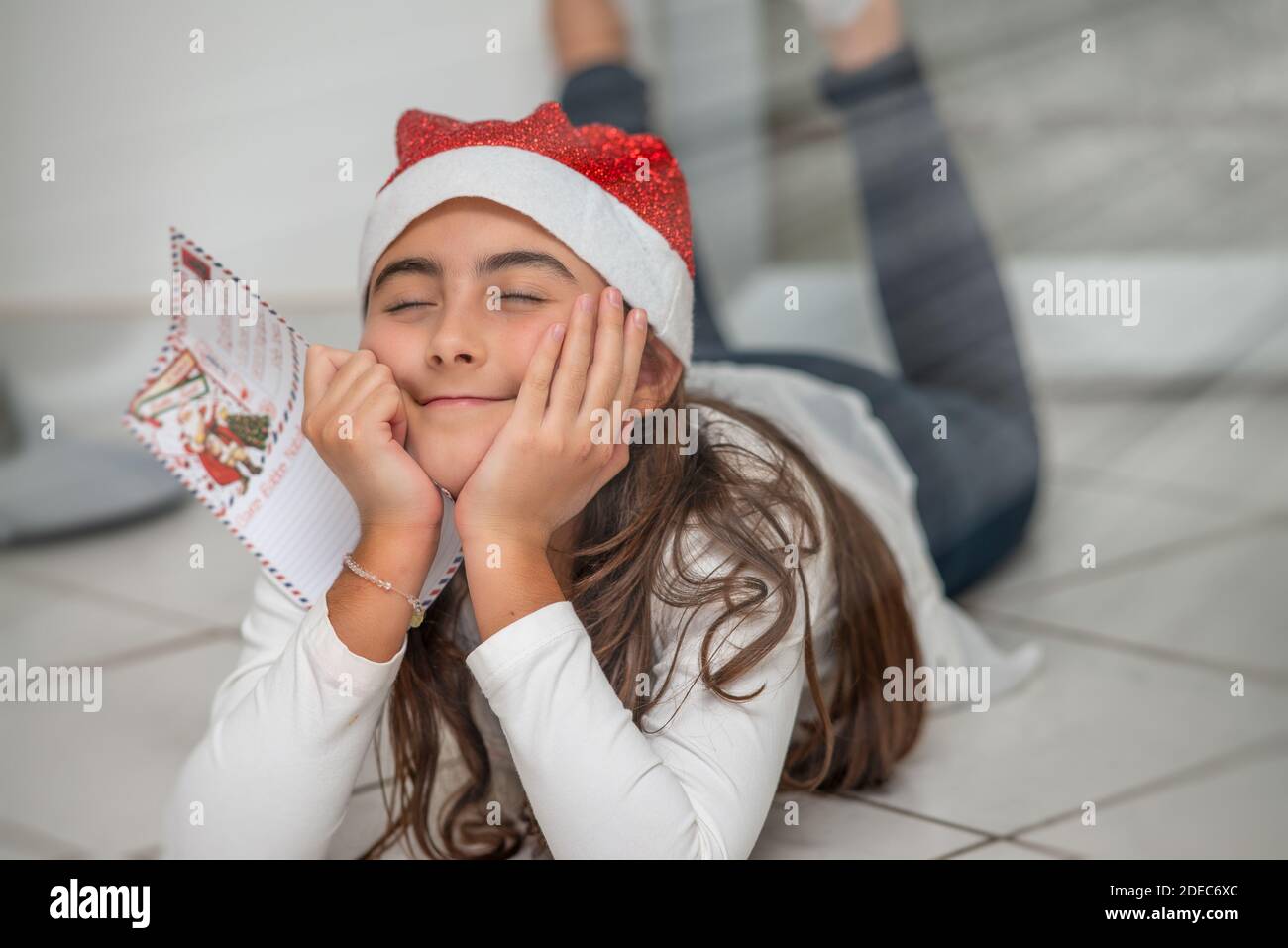 Une jeune fille couchée sur le sol à la maison portant un chapeau rouge de Noël tenant une lettre au Père Noël. Rêver de nouveaux jeux. Banque D'Images
