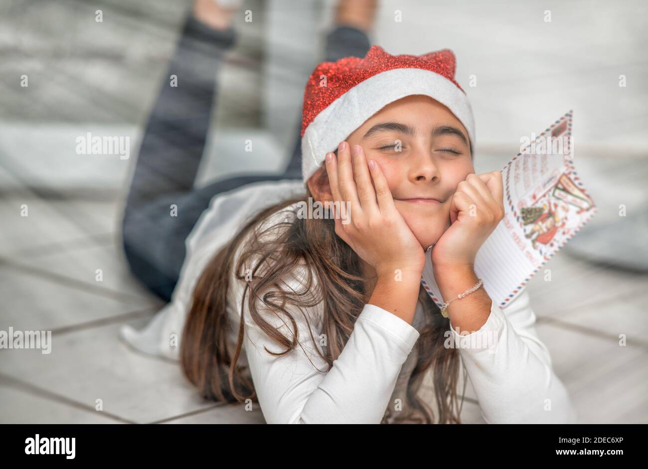 Une jeune fille couchée sur le sol à la maison portant un chapeau rouge de Noël tenant une lettre au Père Noël. Rêver de nouveaux jeux. Banque D'Images
