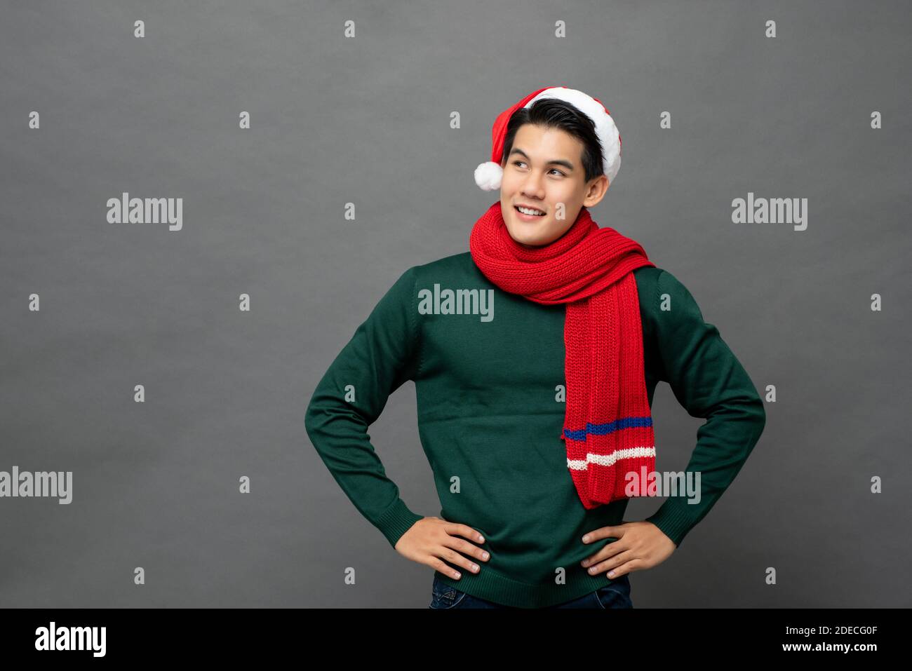 Jeune homme asiatique beau portant des couleurs rouges et vertes de Noël vêtements à thème avec les bras akimbo regardant l'espace de copie de côté isolé sur fond gris Banque D'Images