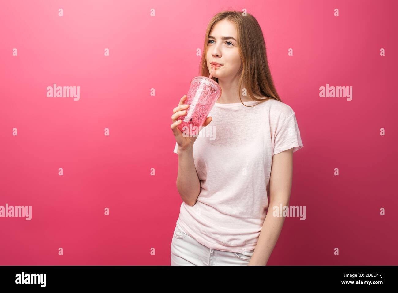 Portrait d'une belle jeune femme buvant une boisson gazeuse dans une bouteille avec une paille et regardant l'appareil photo isolé sur un fond rose, Banque D'Images