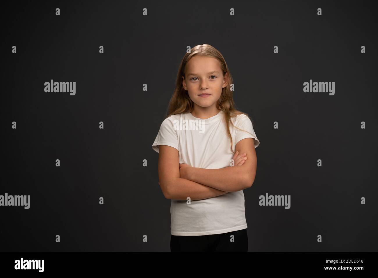 Petite fille de 8,10 ans avec les mains pliées looks questionneusement à l'appareil photo portant un t-shirt blanc isolé sur fond gris foncé ou noir Banque D'Images