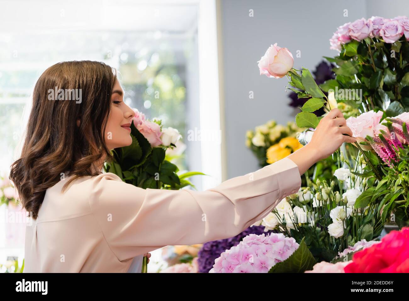 Vue latérale d'un fleuriste souriant prenant la rose en magasin, tout en arrangeant le bouquet avec une fenêtre floue sur l'arrière-plan Banque D'Images