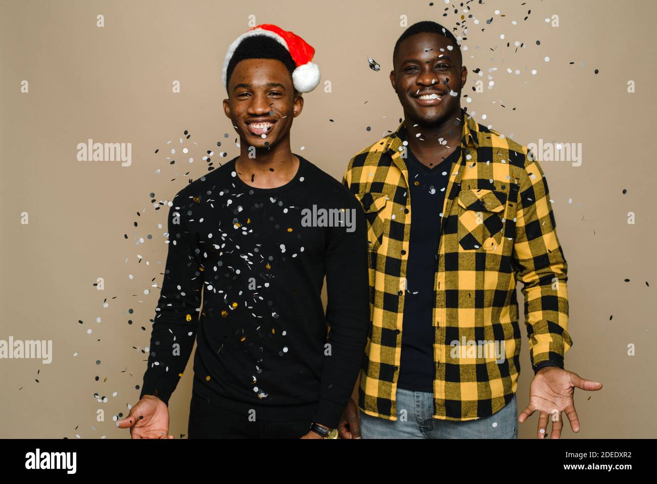 Deux hommes gais noirs élégants et attrayants célèbrent la fête du nouvel an, un couple homosexuel qui jette des confetti, se félicitant mutuellement sur fond beige. Célébration de Noël et du nouvel an. Banque D'Images