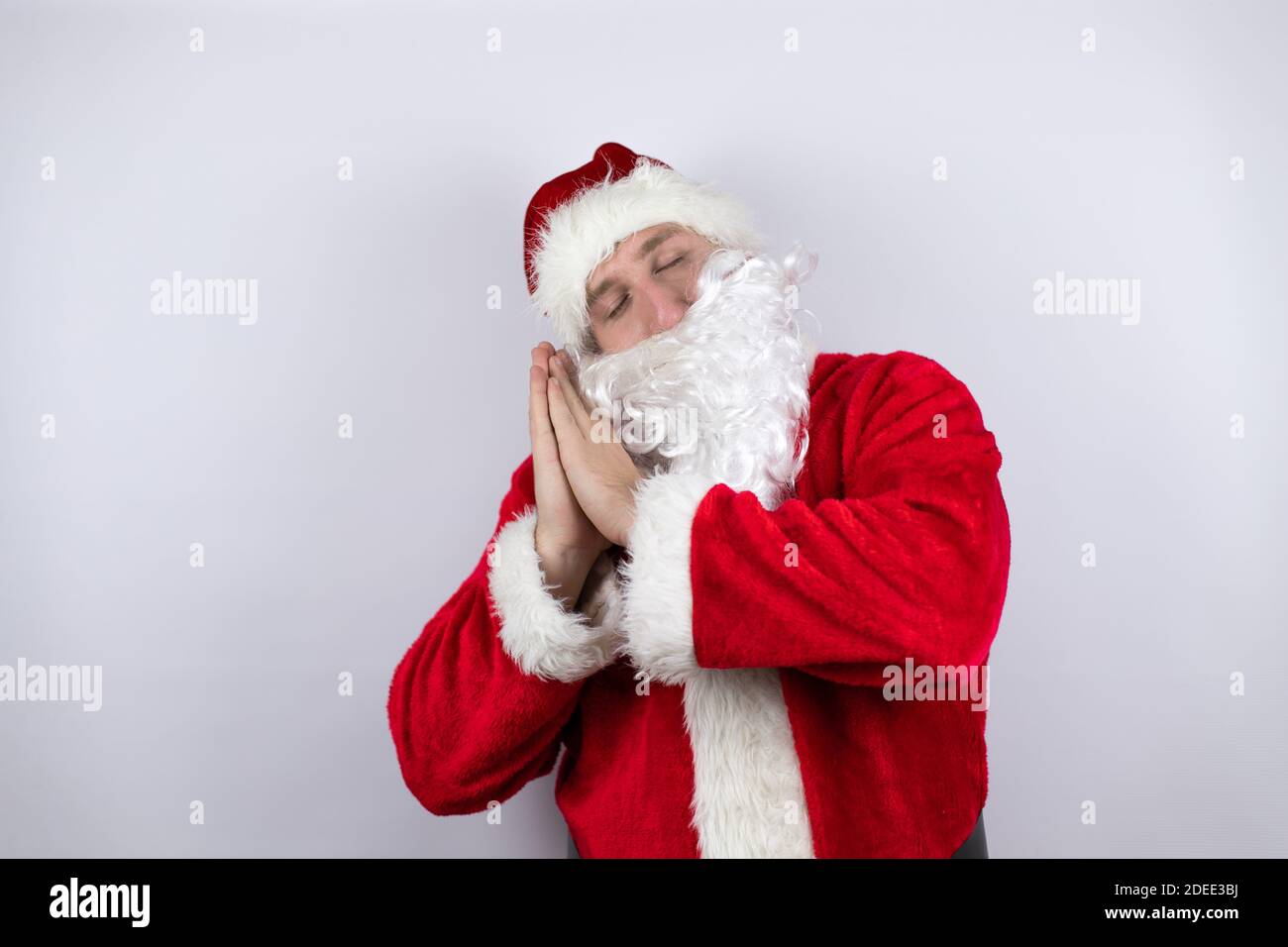 Homme habillé comme le Père Noël debout sur un fond blanc isolé dormir fatigué rêvant et posant avec les mains ensemble tout en souriant avec fermé ey Banque D'Images