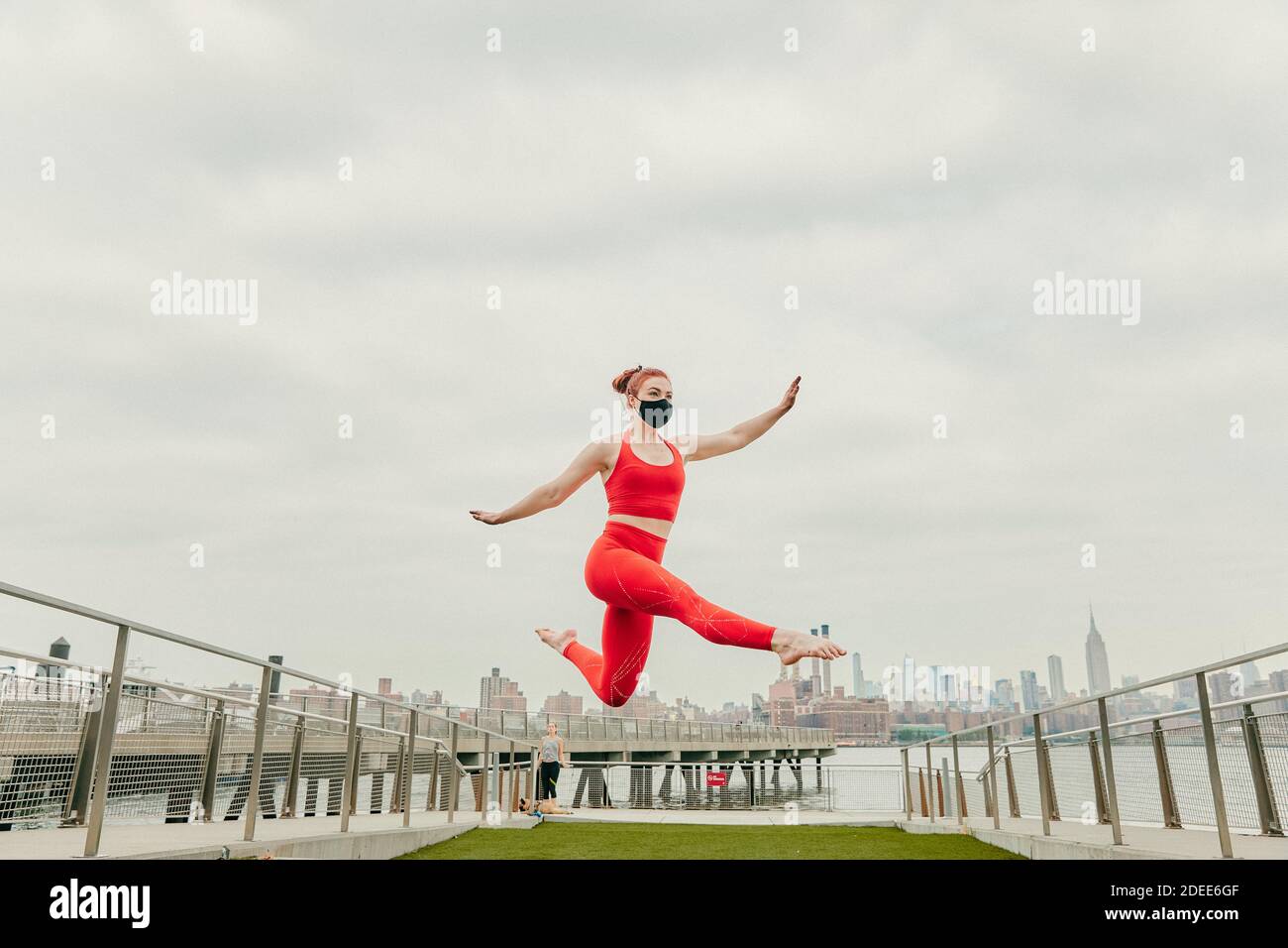 Jeune athlète féminine sautant l'air moyen portant un masque facial par front de mer Banque D'Images