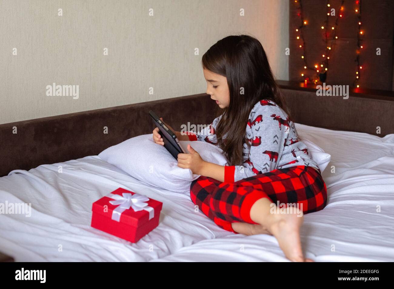 Petite fille de brune en pyjama se trouve sur le lit à la maison, regarde dans la tablette numérique Banque D'Images