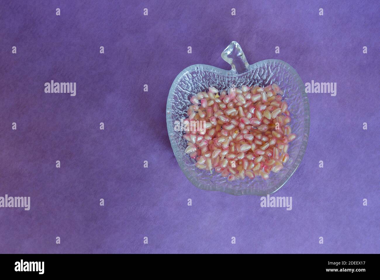 Verre décoratif en forme de fruit contenant quelques graines de grenade prêtes à cuire sur une nappe en tissu peinte en violet. Arrière-plan texturé. Banque D'Images