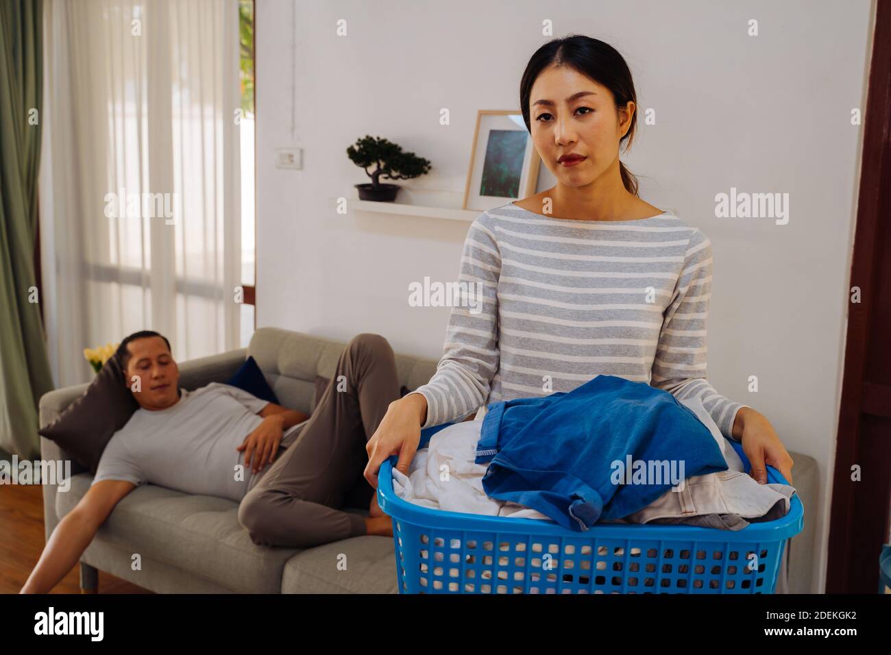 Jeune femme portant un panier de vêtements sales faisant des corvées à la maison tandis que paresseux homme se détendant et dormant sur canapé-lit. Couple ayant un problème domestique de travaux ménagers Banque D'Images
