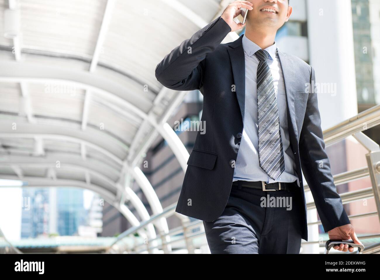 Homme d'affaires qui appelle sur un téléphone portable tout en tirant ses bagages et en marchant dans un passage couvert extérieur - affaires sur la route et concept de déplacement Banque D'Images