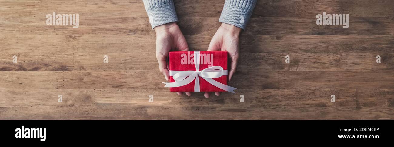 Femme dans un chandail gris donnant un cadeau de noël ou d'anniversaire enveloppé dans du papier rouge et ruban blanc sur une table en bois - vue de dessus, bannière panoramique Web W Banque D'Images