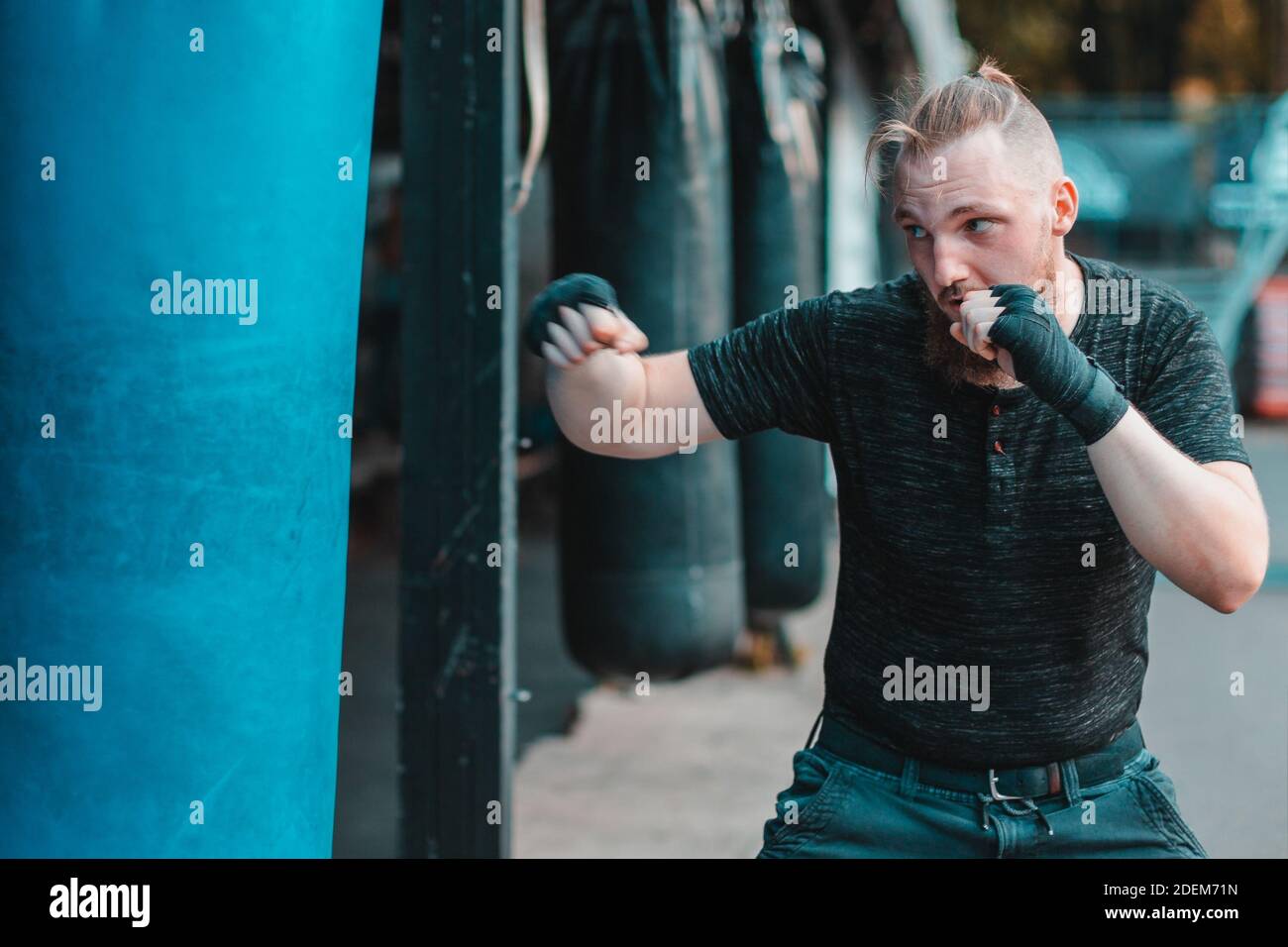 Street Fighter dans les vêtements noirs et les bandages sur le Boxe de poignet dans le sac de Punching à l'extérieur. Jeune homme faisant la boîte d'entraînement et de pratiquer ses coups de poing à la salle de gym extérieure Banque D'Images