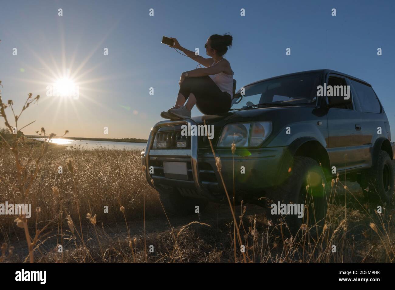 jeune femme prenant un selfie avec son mobile assis le capot de sa voiture tout-terrain sur un lac en été en vacances Banque D'Images