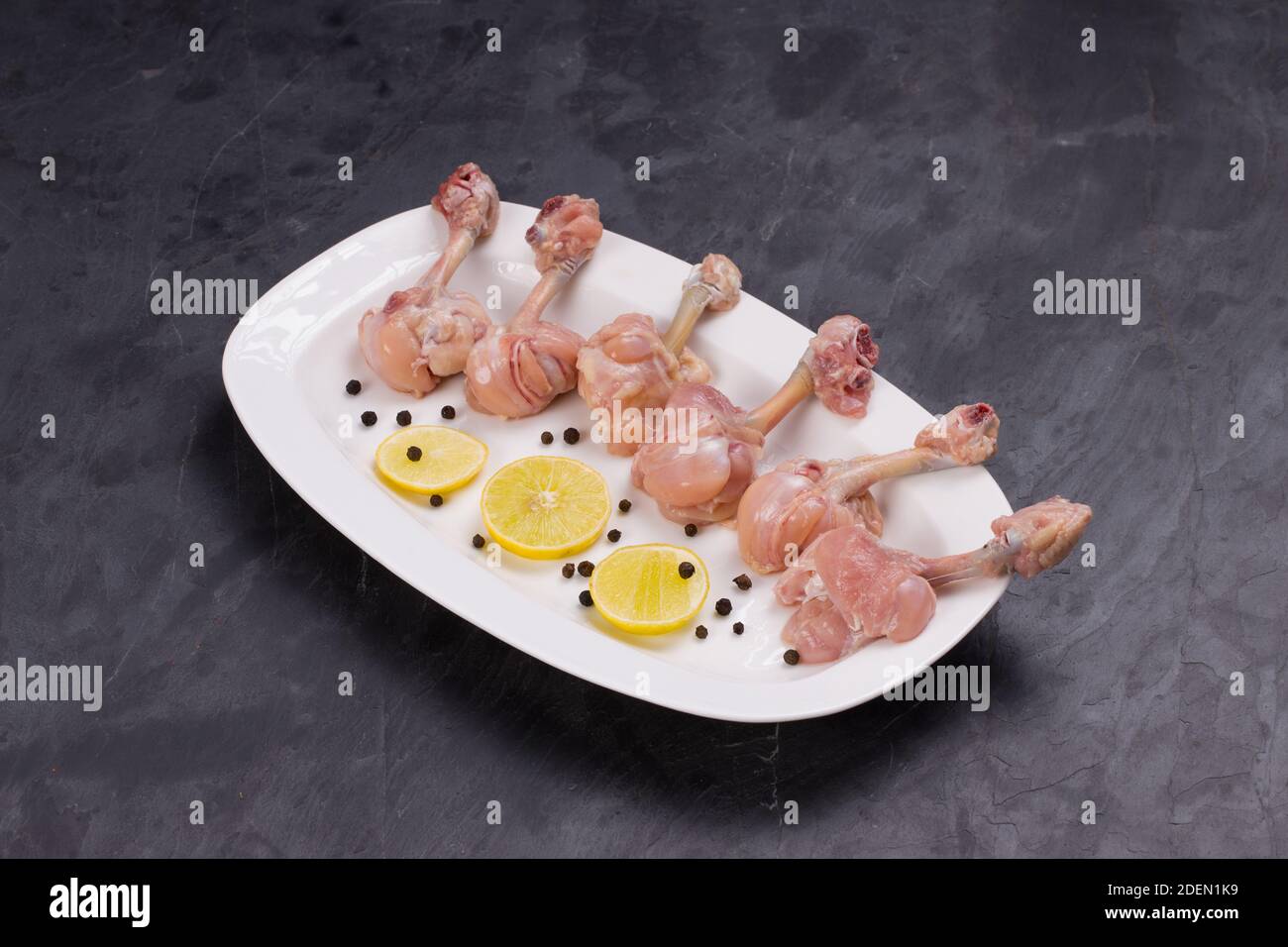 Lollipop de poulet cru, six morceaux de Lollipop de poulet disposés sur un servir une assiette blanche et garni de tranches de citron et de noir poivre sur texture grise Banque D'Images