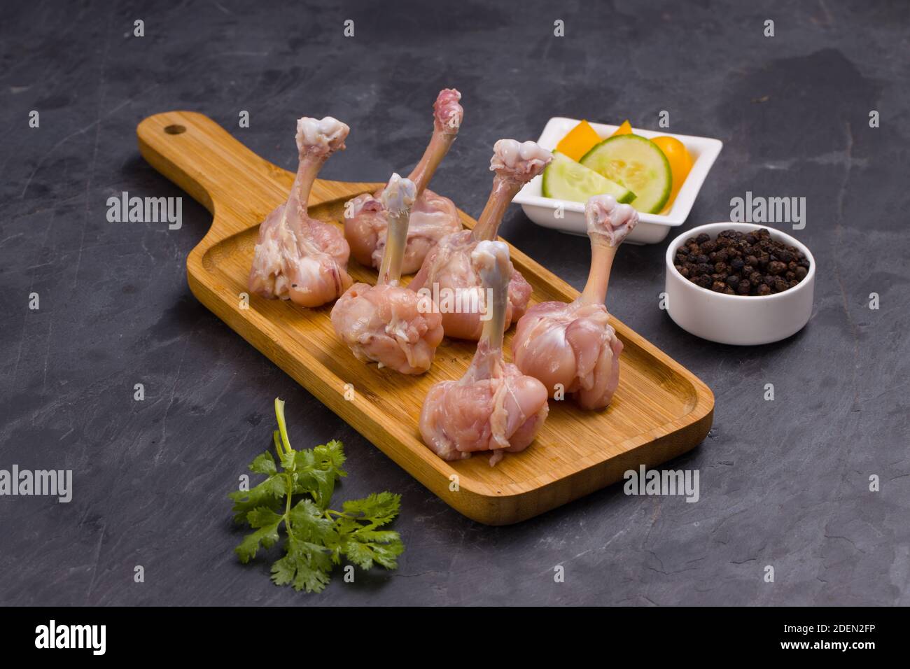 Lollipop de poulet cru, six morceaux de Lollipop de poulet disposés sur un plateau de service avec poivre noir, feuilles de coriandre et tranches de concombre wi en arrière-plan Banque D'Images