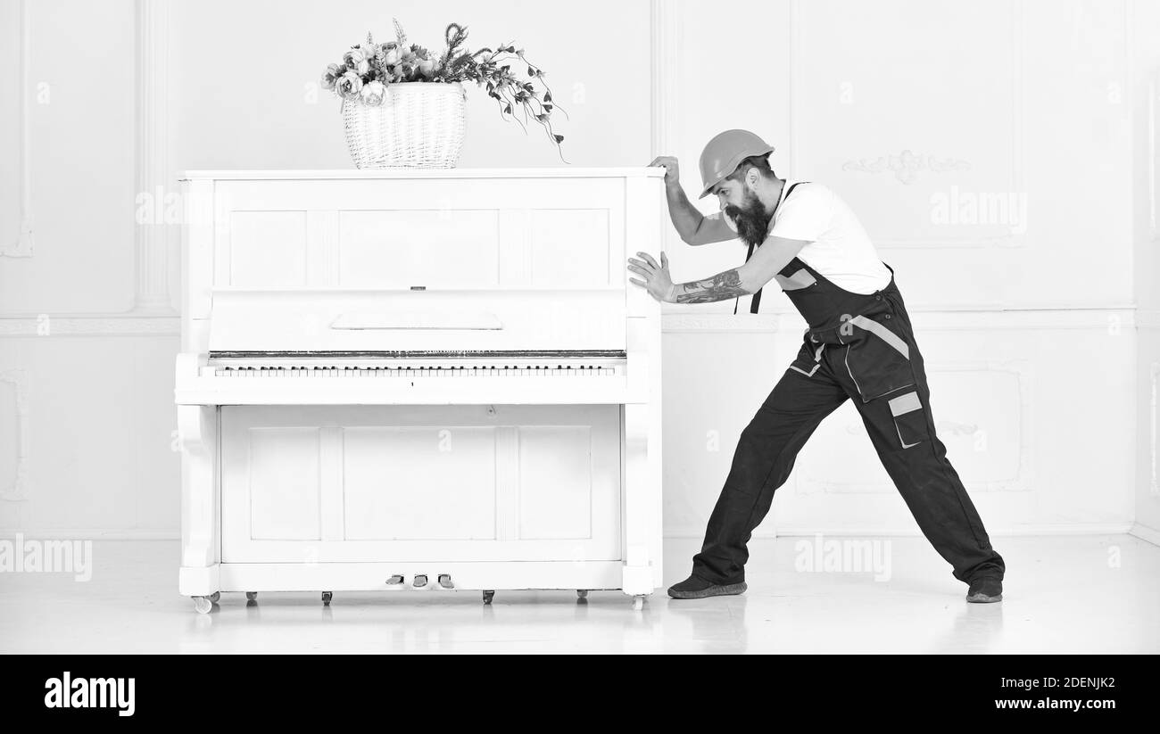 Homme avec la barbe, travailleur en salopette et casque pousse le piano, fond blanc. Le service de messagerie livre les meubles en cas de déménagement. Concept de service de livraison. Le chargeur déplace l'instrument de piano. Banque D'Images