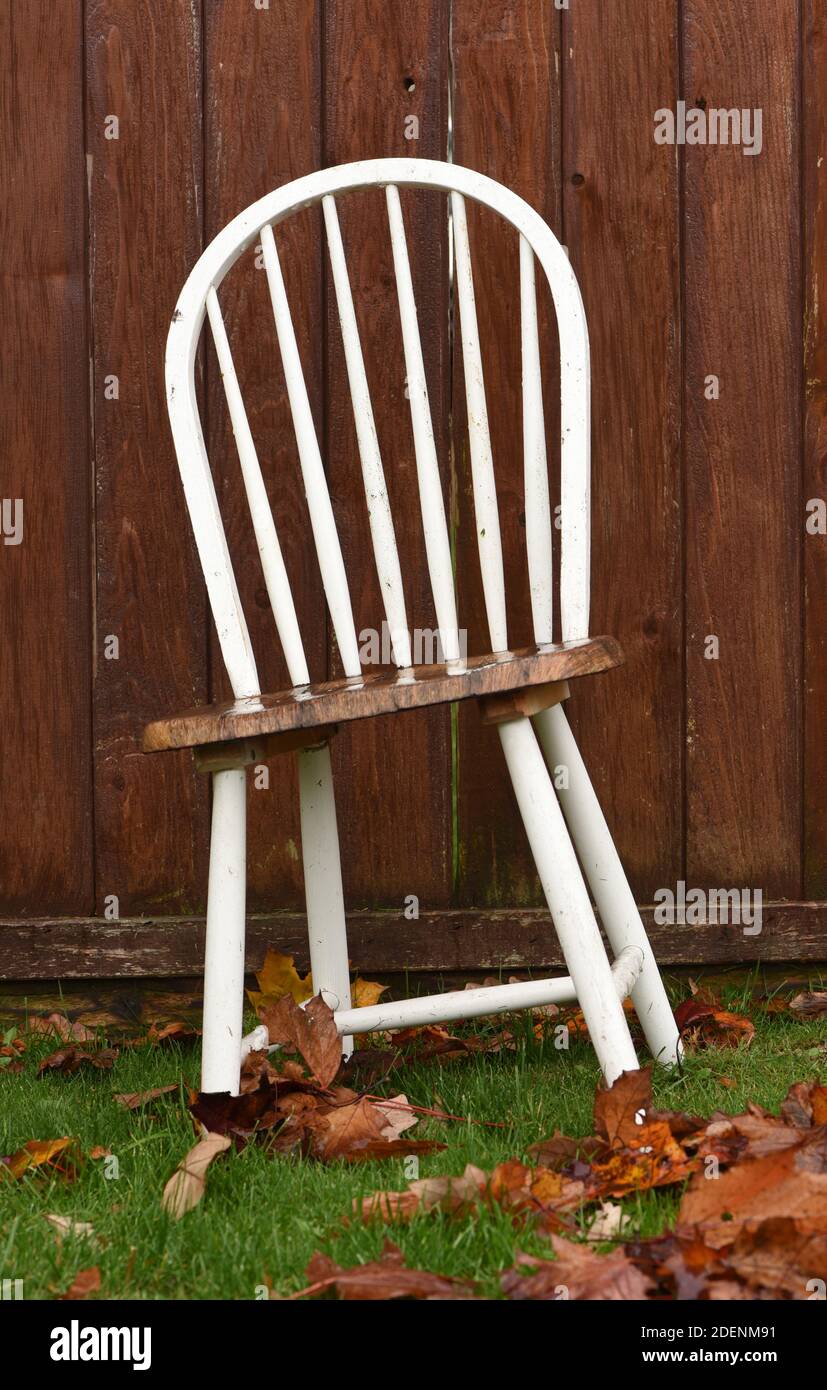 Des feuilles tombées et de l'herbe verte entourent une ancienne chaise en bois peint en blanc appuyé contre une clôture en bois Banque D'Images