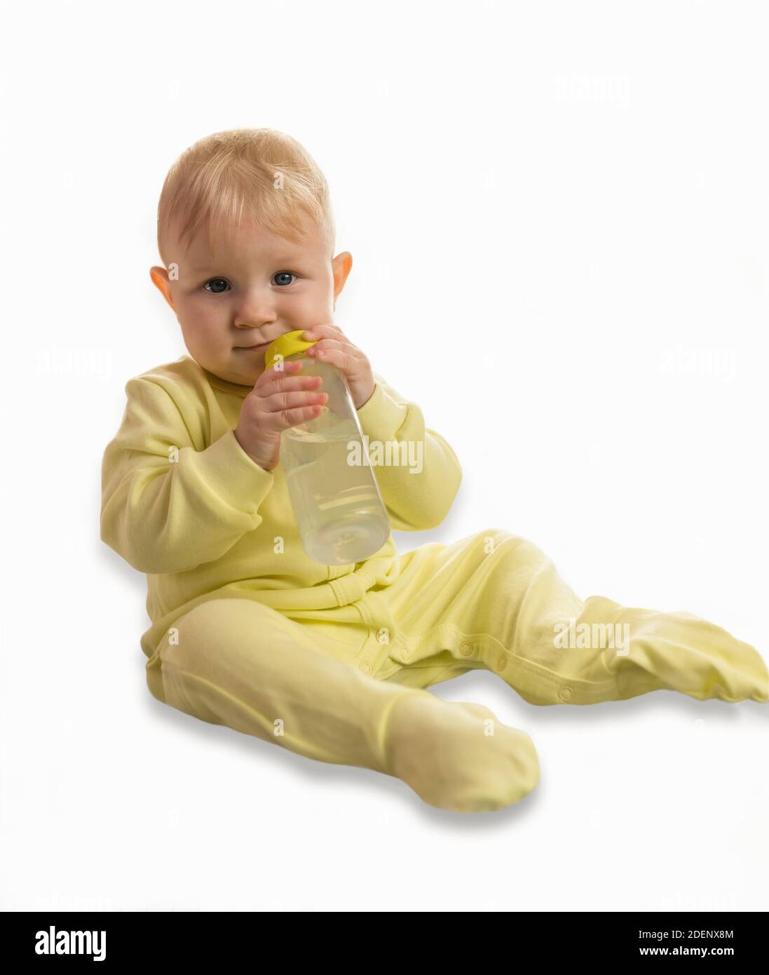 Un bébé avec un sourire dans des vêtements jaunes est assis à la maison avec une bouteille d'eau dans ses mains. Regarder la caméra. Arrière-plan blanc Banque D'Images