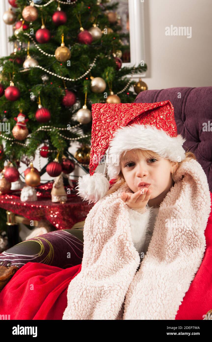 Belle petite fille avec chapeau de père noël enveloppé dans une couverture rouge, assis à côté de l'arbre de christmass et envoyant des baisers Banque D'Images