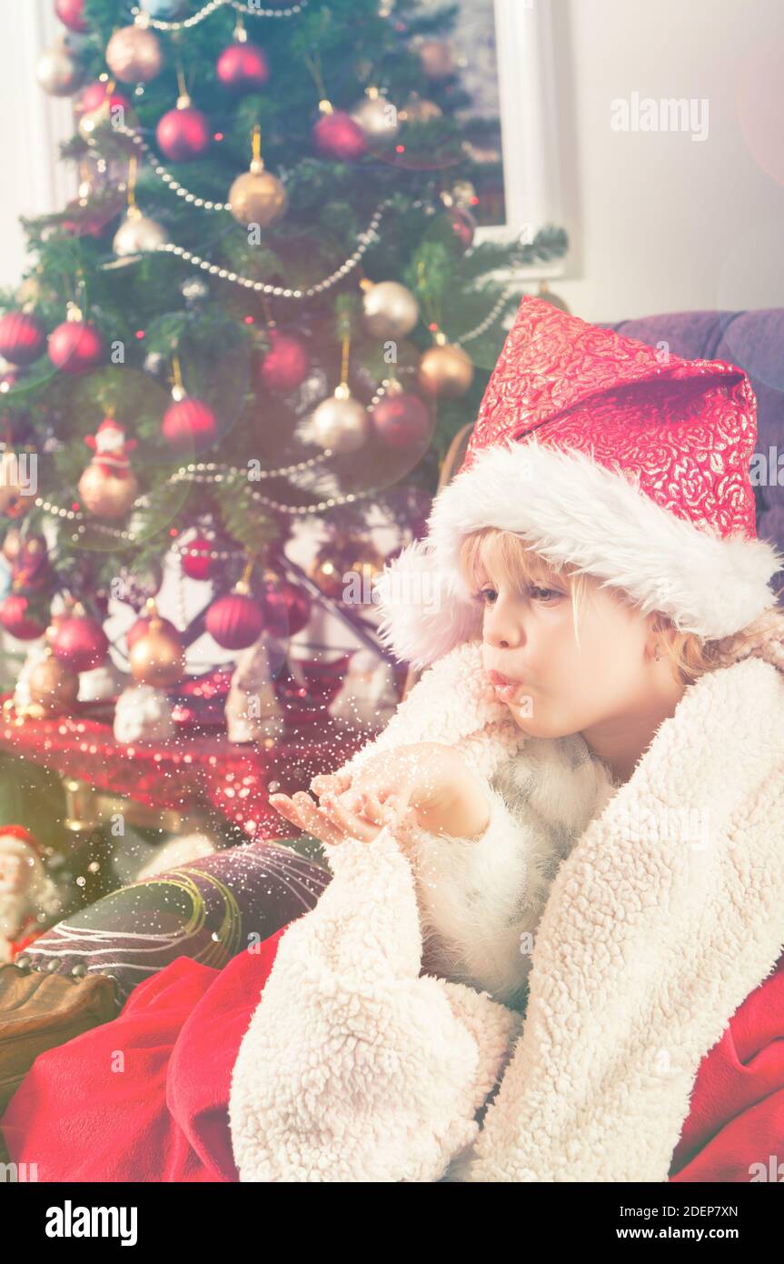 Belle petite fille avec chapeau de père noël enveloppé dans une couverture rouge, assis à côté de l'arbre de christmass et envoyant des baisers Banque D'Images