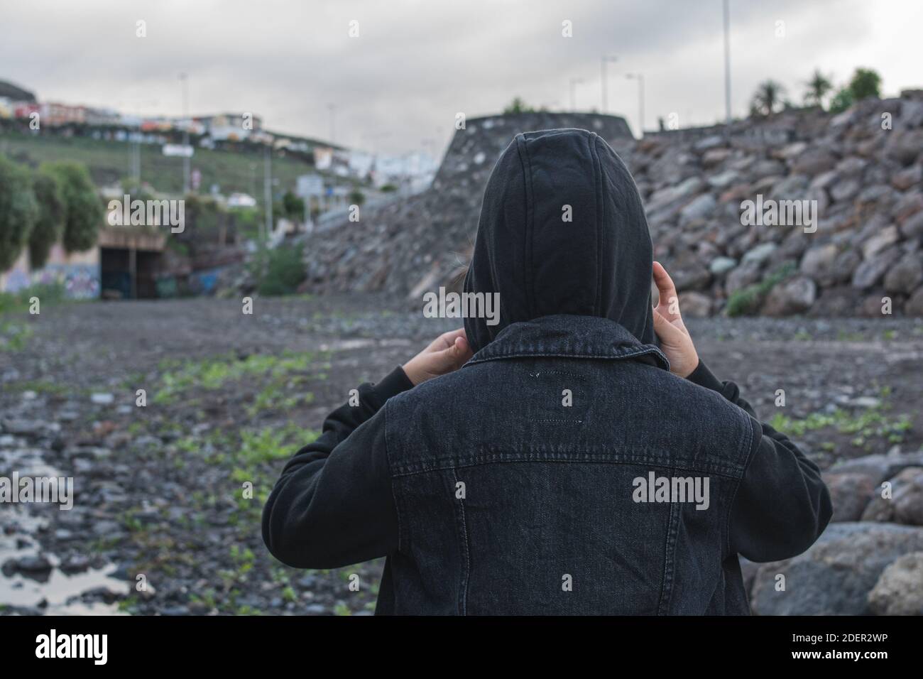 Fille mettant sur la capuche de sa veste noire avec les deux mains, un jour nuageux. Banque D'Images