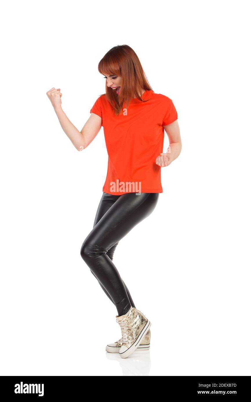 Une jeune femme excitée en t-shirt orange, pantalon en cuir noir et baskets dorées se tient debout, elle se claque et crie. Vue latérale. Longueur totale St Banque D'Images