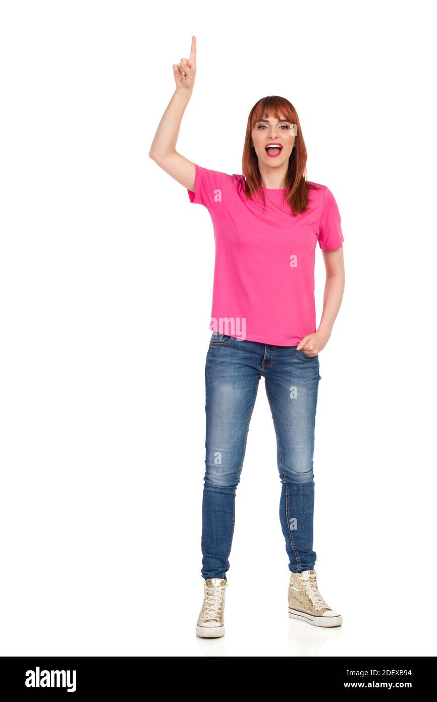 Décontracté jeune femme en chemise rose, jeans et baskets or est debout avec les bras relevés, pointant vers le haut et parlant. Vue avant. La prise de vue en studio pleine longueur est de Banque D'Images
