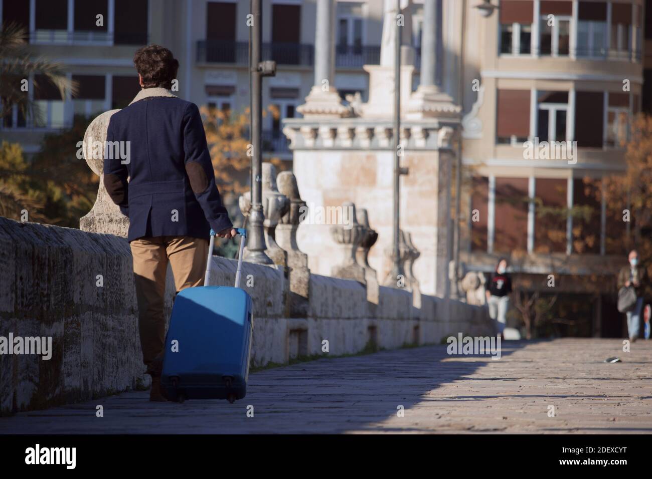 Un jeune homme méconnaissable se rend en Espagne lors d'une pandémie avec une valise bleue tout en marchant dans le centre d'une ville à des fins d'affaires. Banque D'Images