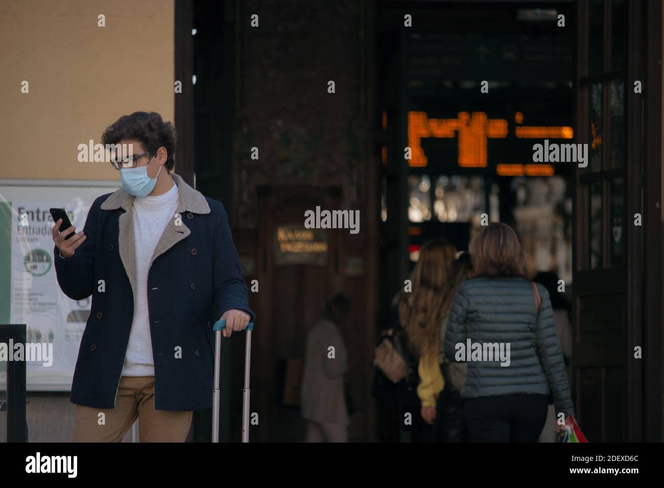 Un jeune homme reconnaissable avec un masque devant la gare avec sa valise appelant un taxi pour le prendre. Voyage d'affaires 2021. Banque D'Images