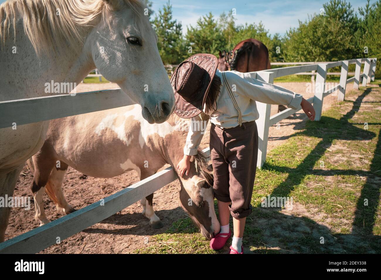 Bonne vie de ferme. Petite fille dans un stand de tenue vintage près du paddock avec un beau cheval blanc et petting un poney. La fille est en parie Banque D'Images