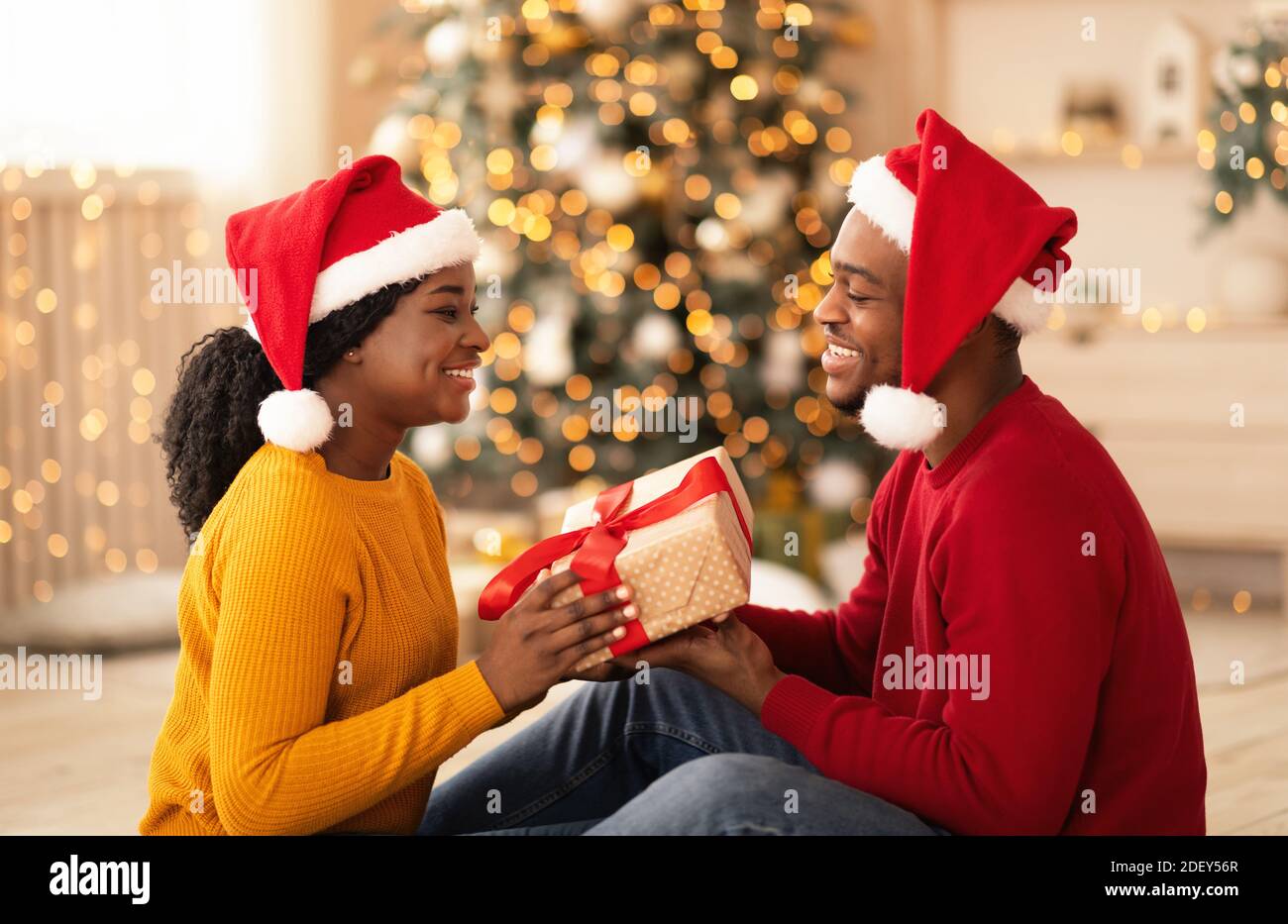 Joyeux jeune africain américain homme et femme en chapeaux de Santa échangez des cadeaux à l'intérieur Banque D'Images